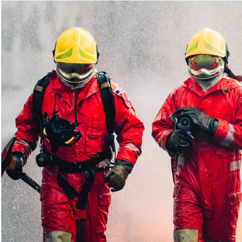 Two men in red fire suits walk through a spray of water, showcasing their readiness and teamwork in a firefighting scenario.